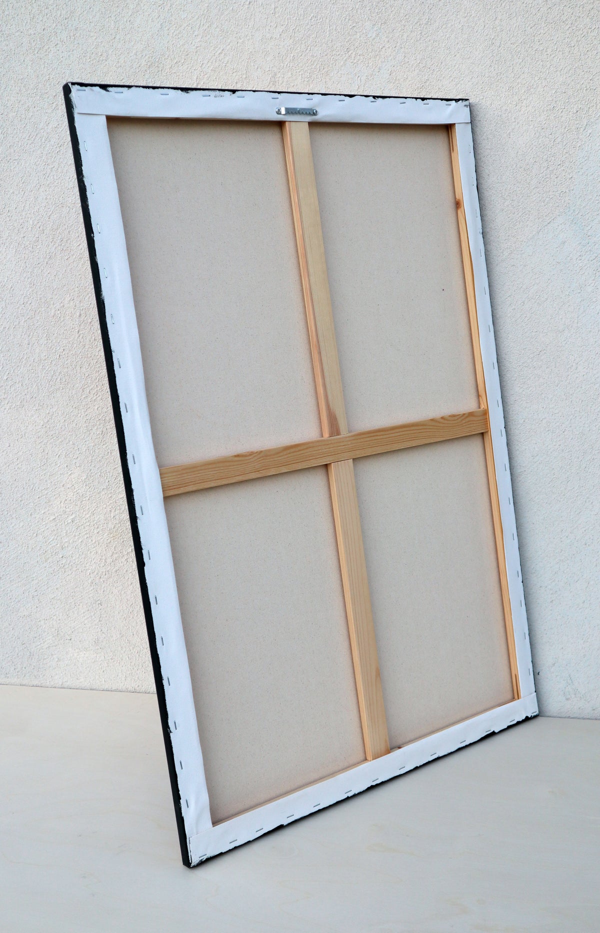 Back view of a 24" x 36" canvas, stretched on wooden stretchers with two wooden cross pieces, seen at a 3/4 angle against a light beige wall and floor. 